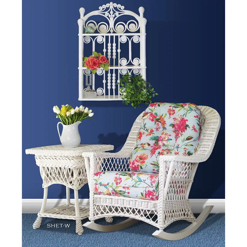 White wicker rocker with floral cushions beside a matching end table holding a pitcher of tulips, under a white wall shelf