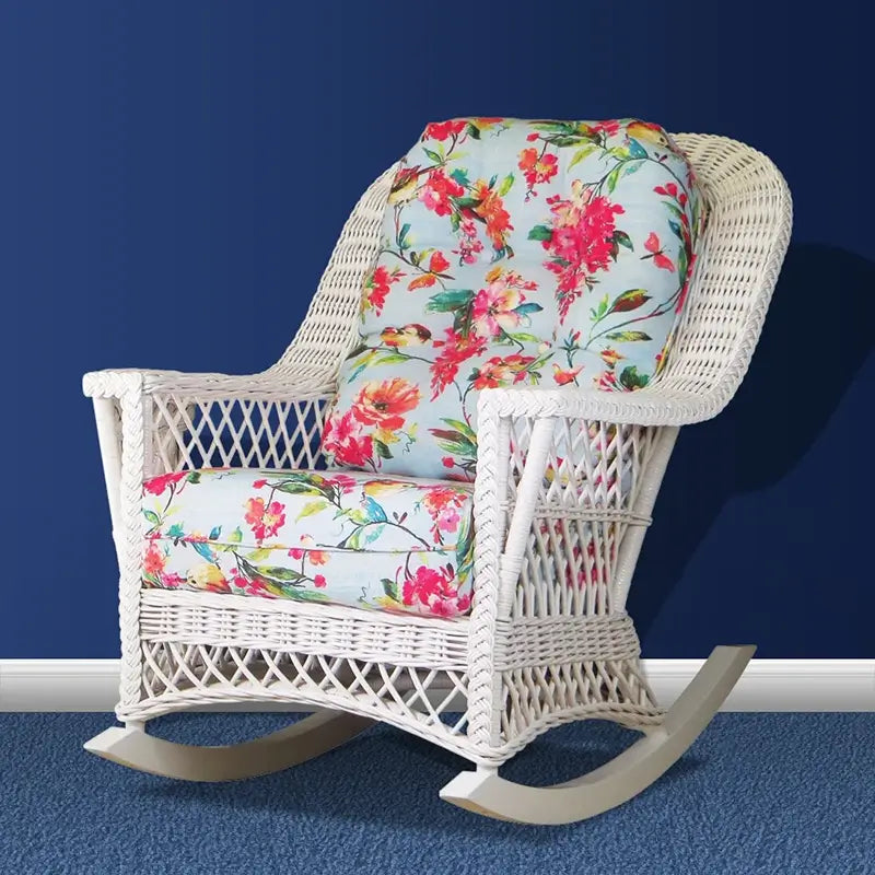White wicker rocking chair with floral seat and back cushions in pink, red and aqua, set against a navy blue wall
