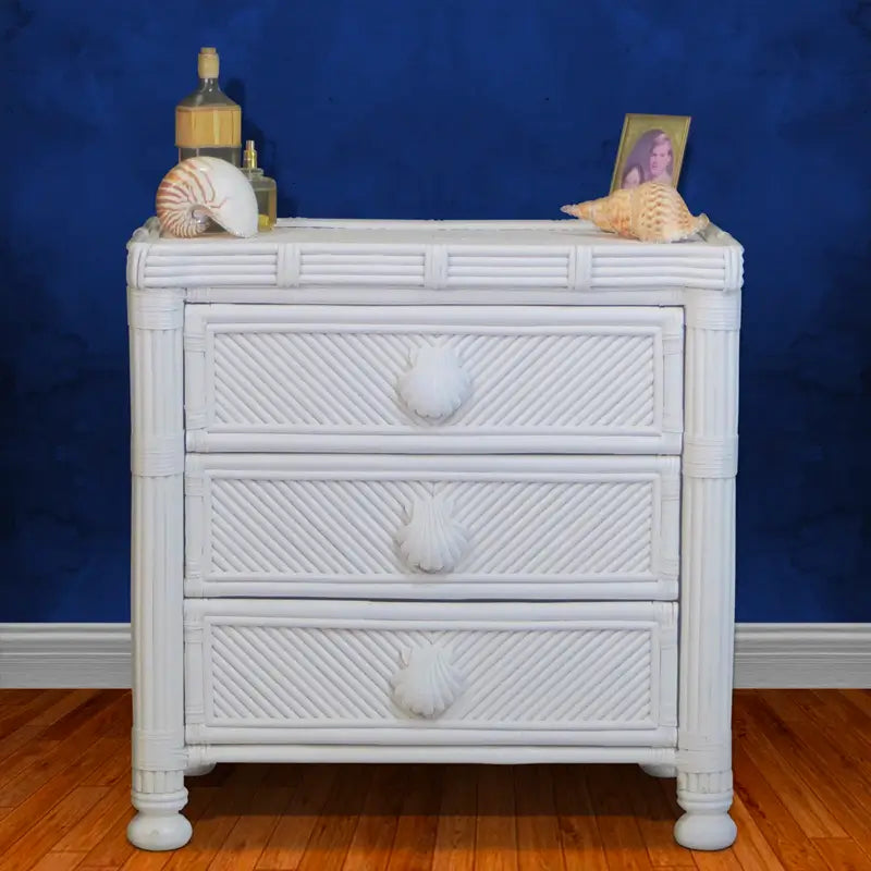 Small white rattan dresser with seashell pulls against a navy blue wall, topped with seashells and a picture frame