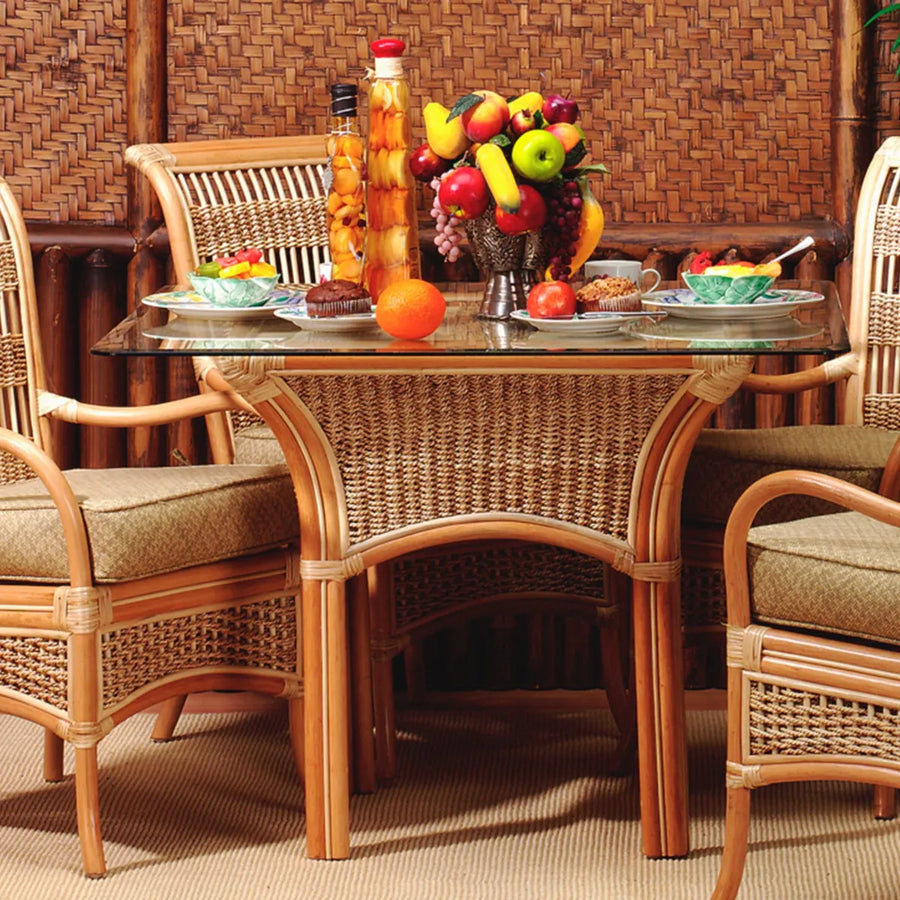 Glass top table from the Spice Island 5 piece rattan dining set in natural, set with fruit and plates, with cushioned arm chairs pulled up around it
